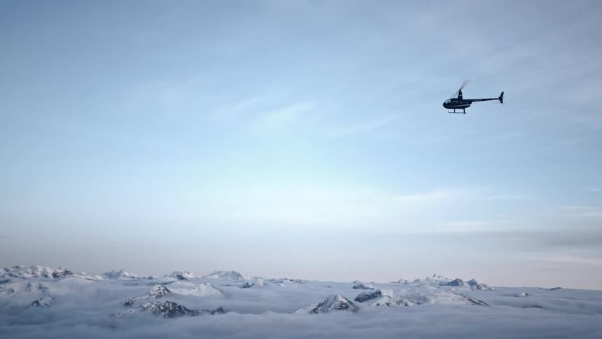 A helicopter flies in clear skies over a snowy and rocky terrain during the day