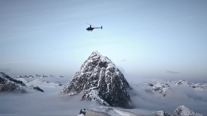 Helicopter flies over a mountain summit lightly covered in snow at daytime