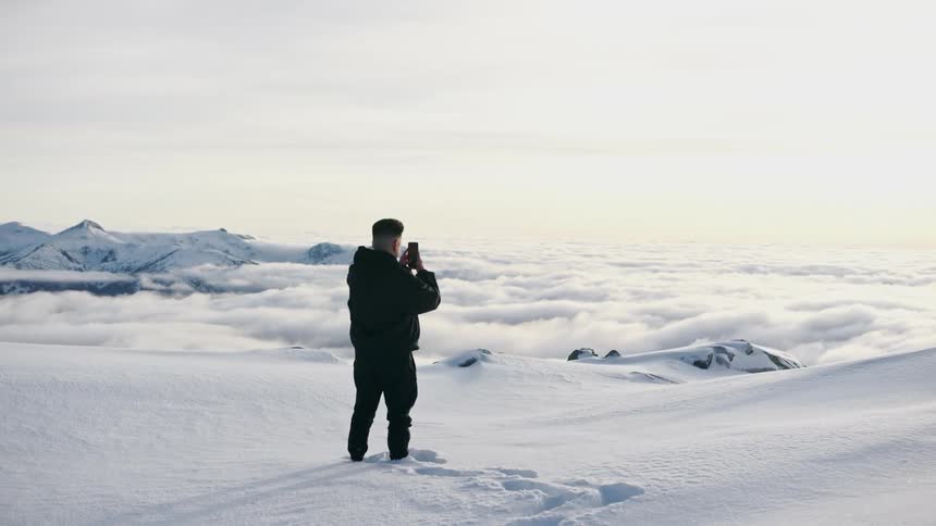 A man stands in snow up to his ankles and takes a photo on his phone of the horizon