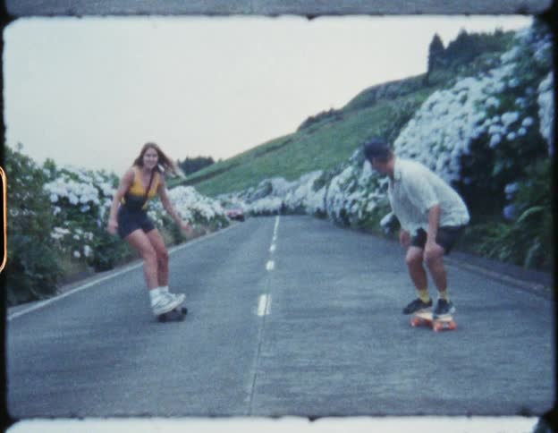A teenage girl skateboards down a flower lined road with her dad
