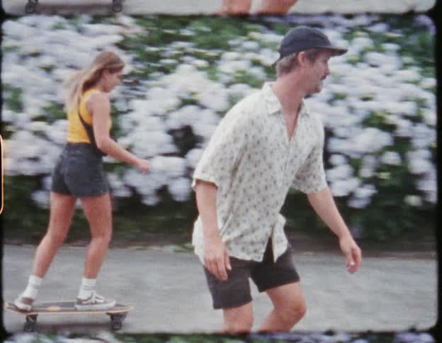 Two friends riding skateboards on a road surrounded by flowers