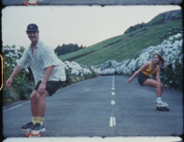 A man and a woman on skateboards rolling down a street in between white floral bushes