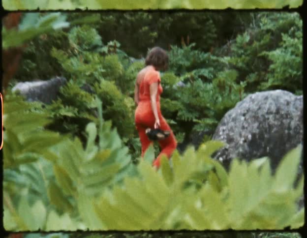 A woman in an orange jumpsuit holds her shoes as she walks barefoot over rocks in a jungle