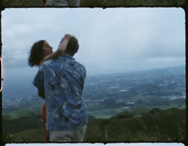 A man and a woman touch one another and hug each other on a hill overlooking a suburban area