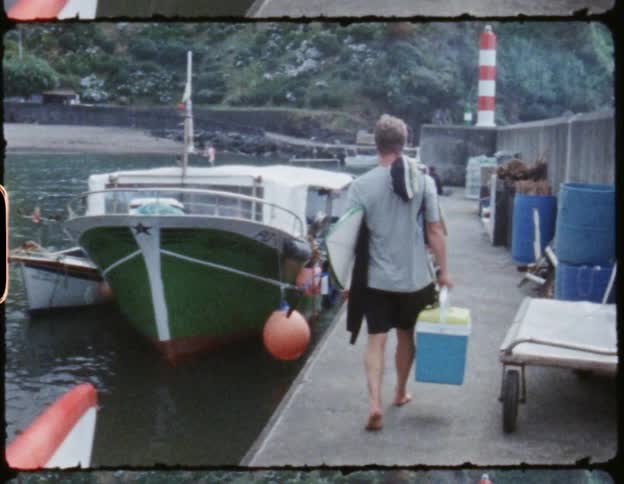 A man holding a cooler and surfboard is walking on a dock toward a boat