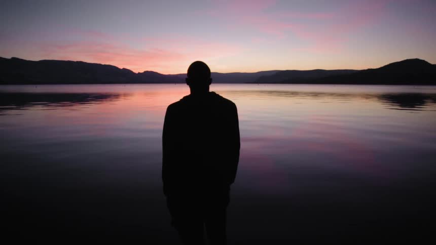 A man stands near a lake at dusk