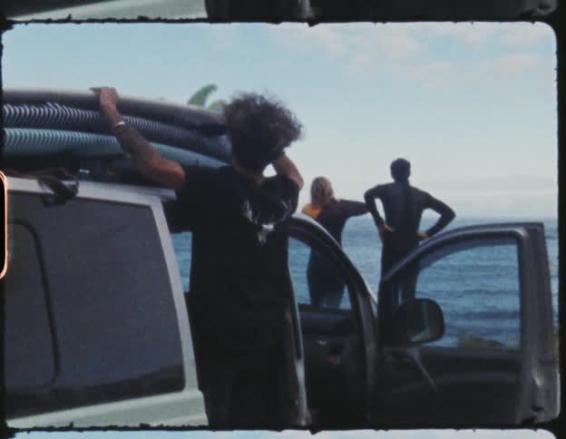 A man takes a surfboard off the top of a vehicle parked behind two other people facing an ocean