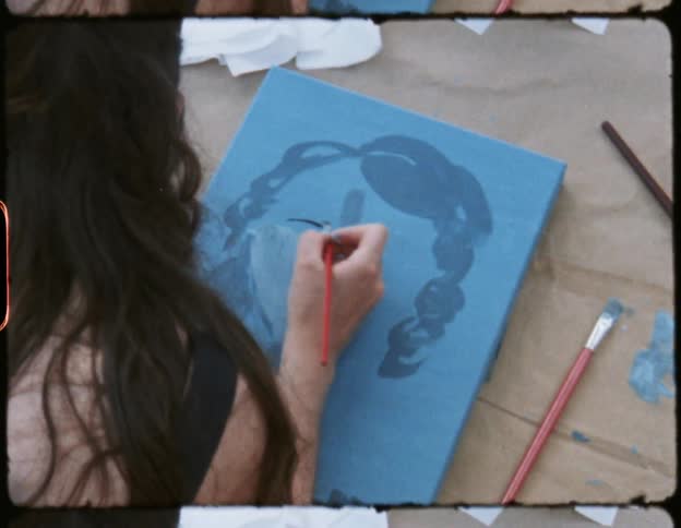 A woman paints a portrait while sitting outside on the ground