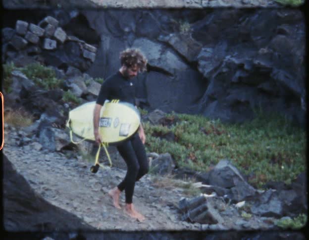 Surfer wearing a black wet suit carries a yellow surfboard down the slope of a beach