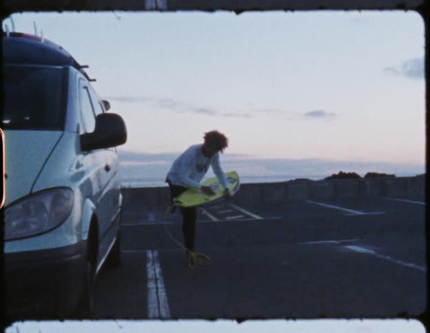 A man uses a cloth to scrub the surface of a surfboard while standing next to a vehicle in a parking lot overlooking the coast during the day