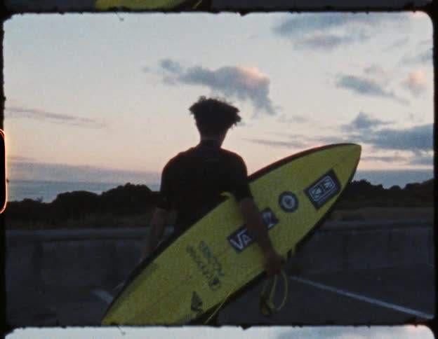Man walks barefoot across parking lot carrying surfboard toward slightly pink tinted sky