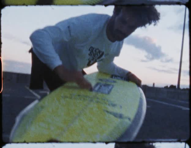 Man standing in a parking lot waxing a yellow surfboard in the evening