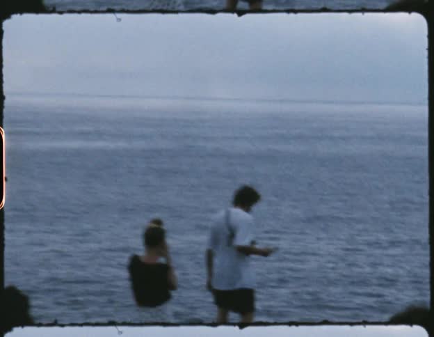 A woman and man stand outside overlooking an ocean with a man walking behind them