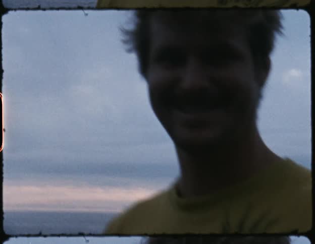 Man smiling at a camera outside next to a woman who is staring off at the ocean at the edge of a cliff