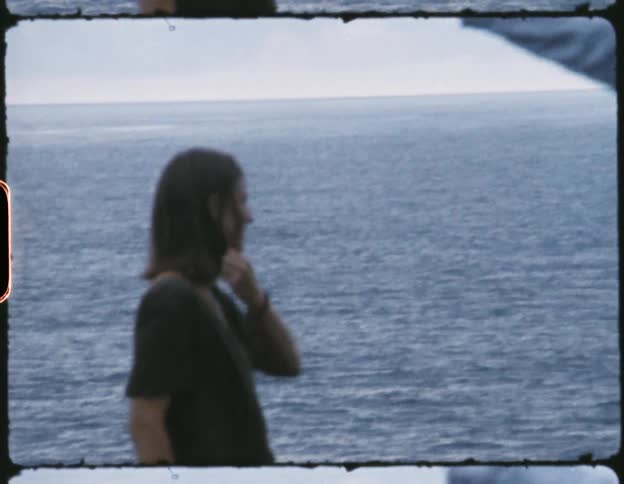 A woman pushes hair out of her hair as she is joined by another person to overlook the ocean