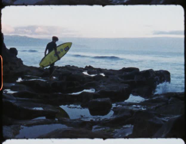 Man walking over rocks next to the ocean carrying a yellow surfboard