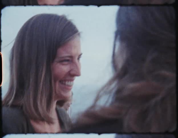 Two women stand in front of a body of water outdoors smiling and laughing