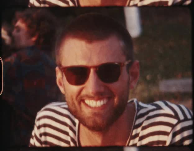 A male smiling surrounded by a few people on a grass lawn