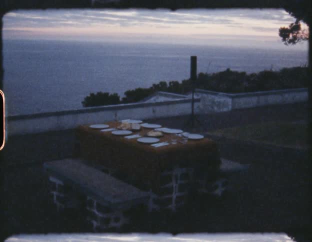 Picnic table with place setting sits near wall overlooking ocean as horizon shines pink