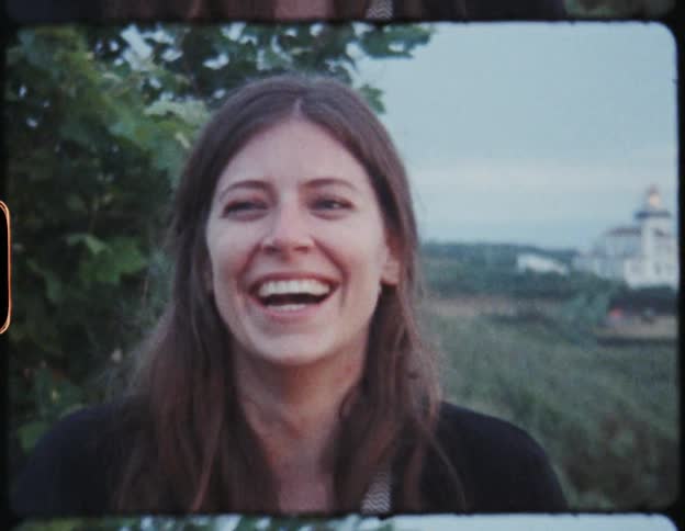 A young woman laughs while talking pictures with a large camera alongside a group of people outdoors during the day