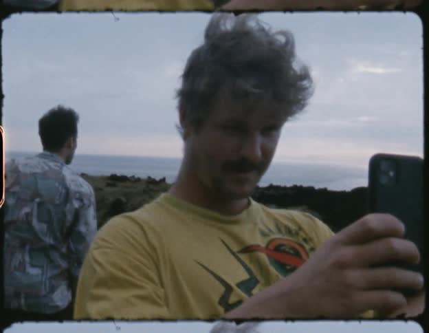 Man comes into view from in front of a cliff side overlooking the ocean and uses his smartphone to record the camera