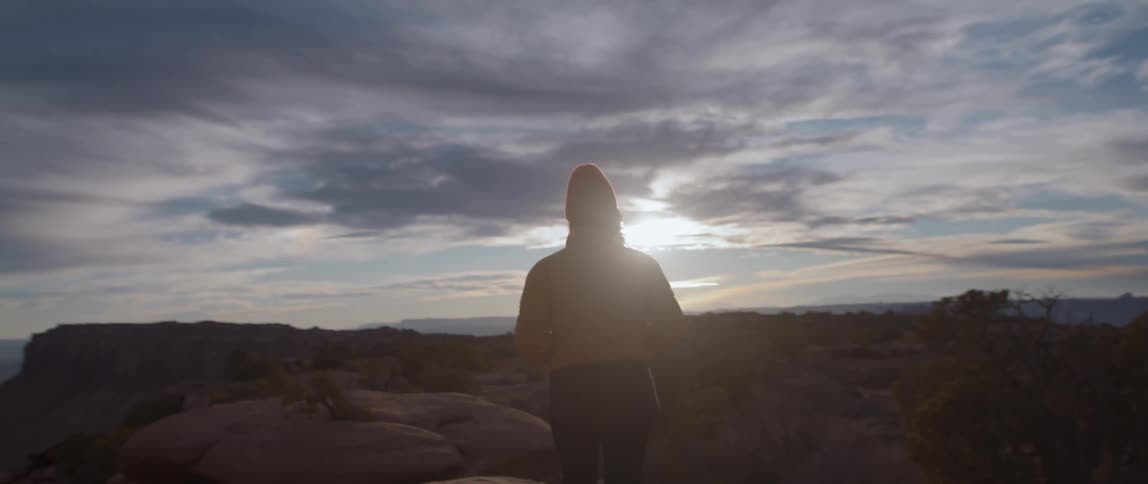 A woman walks across a plateau in a canyon with her hands tucked into her yellow coat