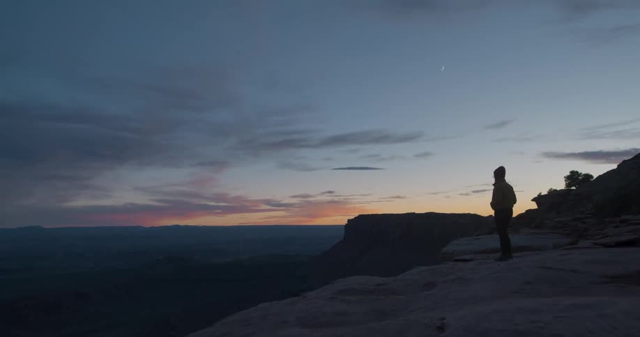 A person walks to the edge of a tall mountain and looks at the night sky
