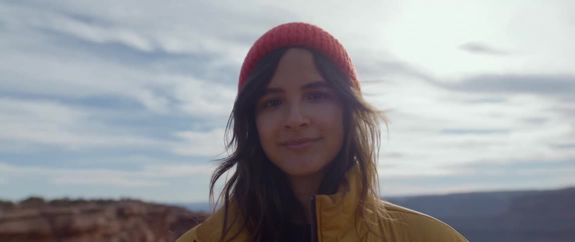 A woman reading a yellow jacket and red beanie smiling at the camera outdoors next to a mountain range