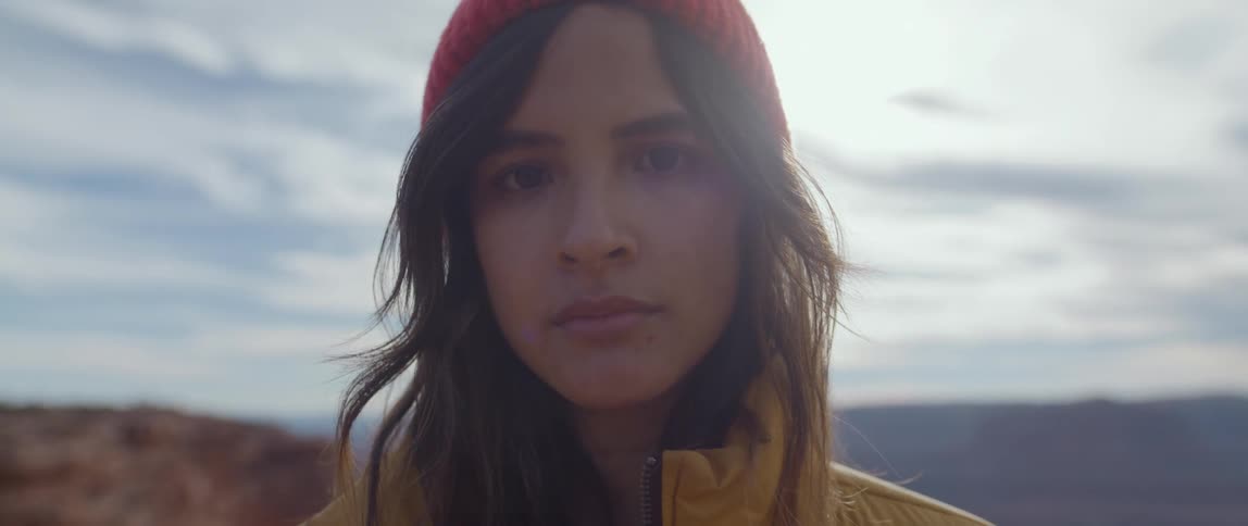 A young woman wearing a heavyweight jacket stands outdoors while looking directly at the camera