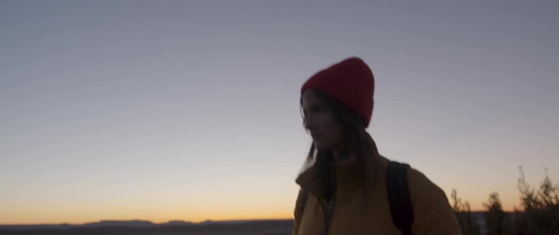 Woman walking outdoors wearing cold weather attire next to a sunset sky