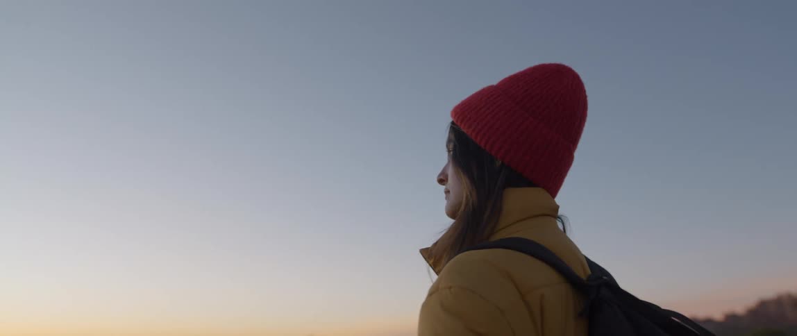 A female hiker wears a hat and coat as she stands outside and looks across the horizon