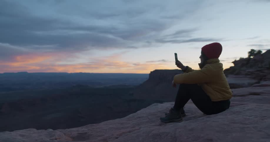Woman sitting atop a mountain takes pictures with her smartphone at sunset