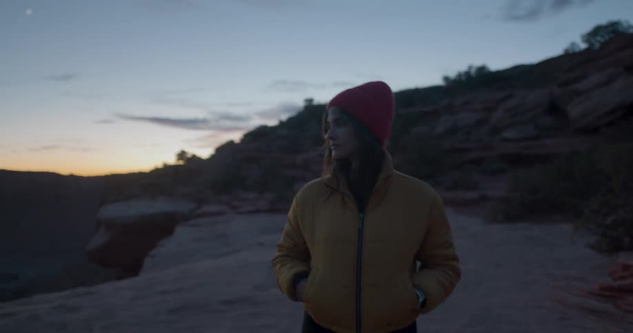 A woman with a hat and a jacket walking in the desert with the sky and clouds in the background