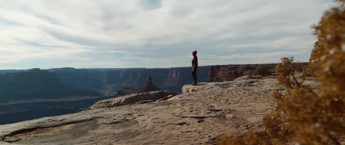 Person stands on edge of cliff looking across misty valley under clouded sky