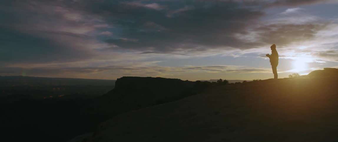 A woman walks across the top of a canyon and stops to take a picture at sunset