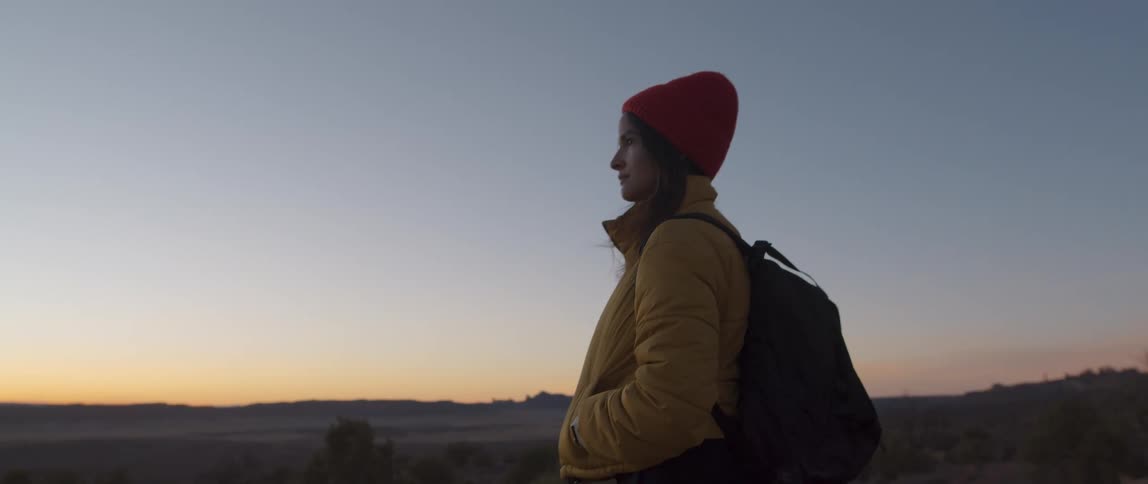 A woman with a backpack a hat and a jacket standing in the desert with the sky and clouds in the background