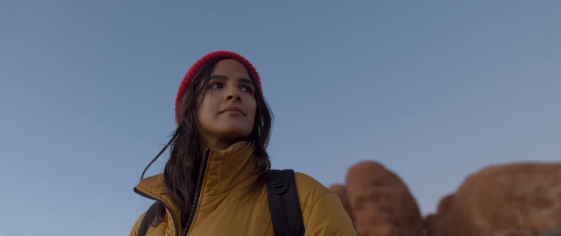 A woman is standing in front of a large rock wearing a hat jacket and backpack during the day with clear skies
