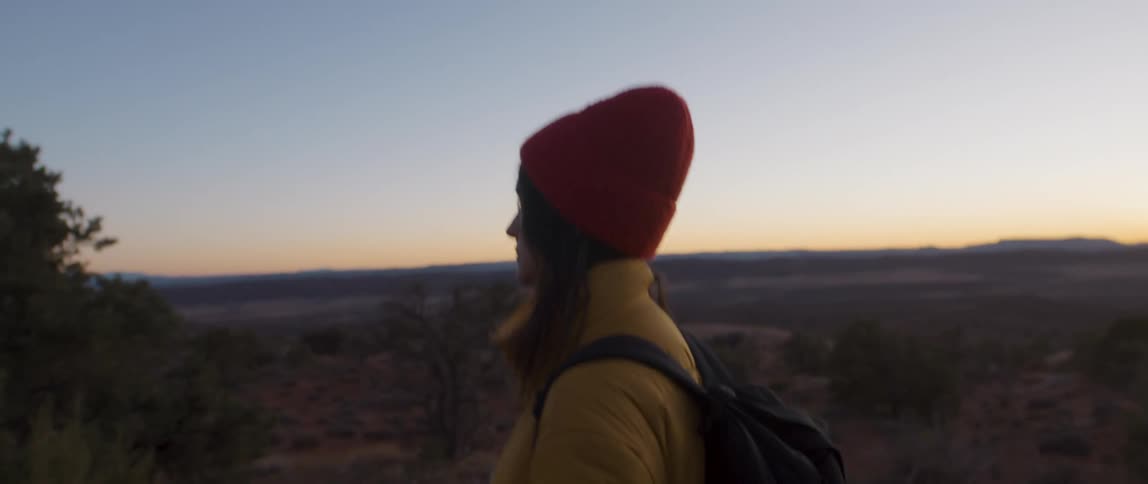 A woman walks by herself in a rural grassland area wearing a hat and backpack