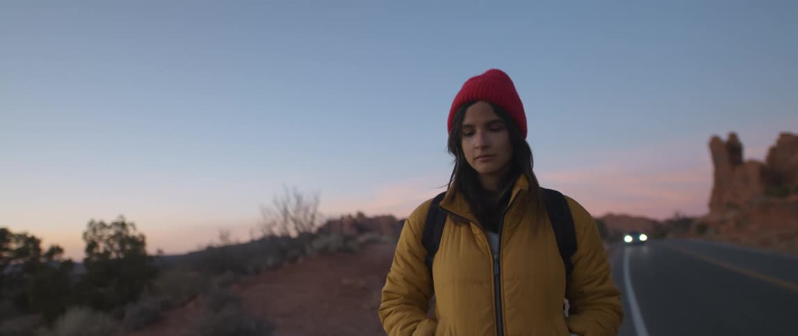 A teenage girl walks along a highway as a car drives up behind her