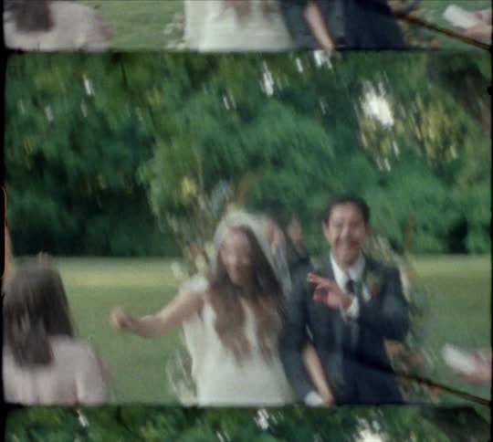Bride and groom walk down an aisle at an outdoor wedding waving at people around them