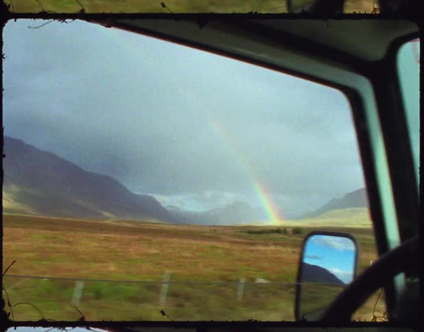 A vehicle is driving and a rainbow has appeared in the grassland to the left of the vehicle