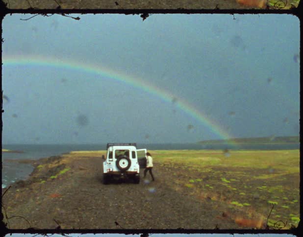 Rain falling causes a person to run for cover in their vehicle as a rainbow spans the cloudy sky