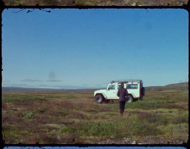 A woman walks towards a parked white car on a grassy field