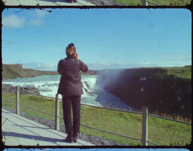 A woman walking on a path next to a waterfall stops and takes a photograph then keeps walking