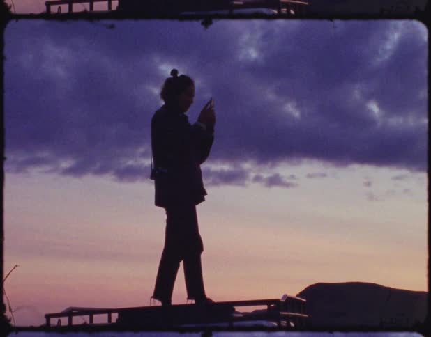 A person stand on top of a vehicle texting on phone with clouds in the sky