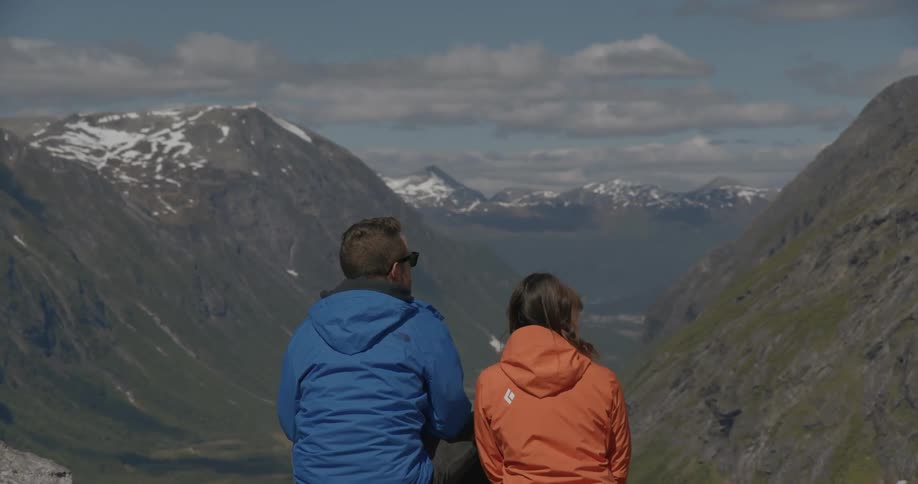 A couple sit and look out at a vast valley between snow-covered mountain peaks