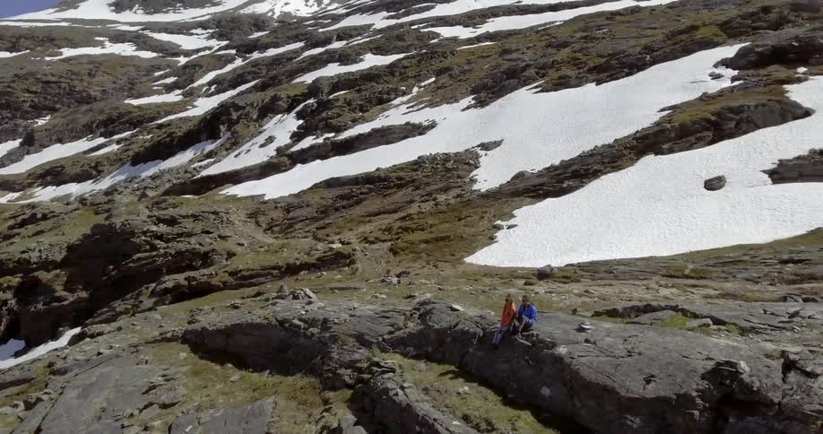 A man and woman sit on a rock ledge on a mountain covered with patches of snow