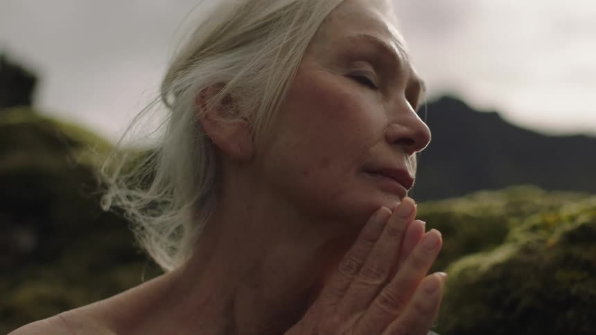 Senior woman with white hair stands in the mountains with praying hands