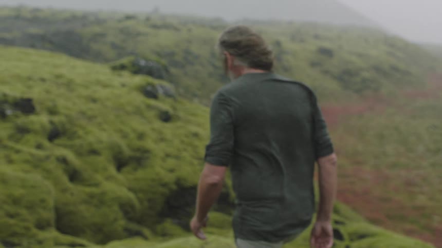 A long haired man walking over the green hills on a windy day