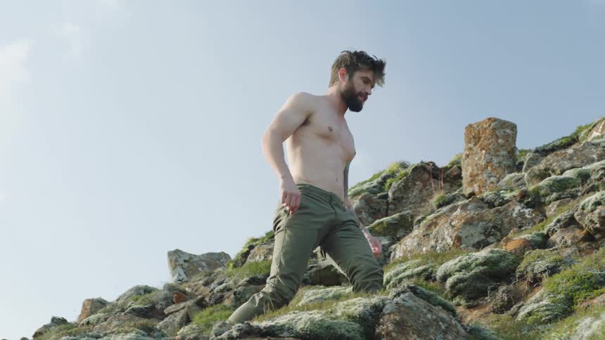 Young bearded shirtless man climbs barefoot the moss covered rocks on a hill